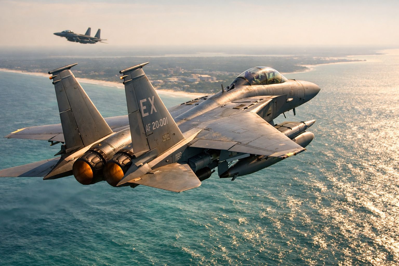 Dois caças militares a jato a voar sobre o mar com uma costa ao fundo ao pôr do sol.
