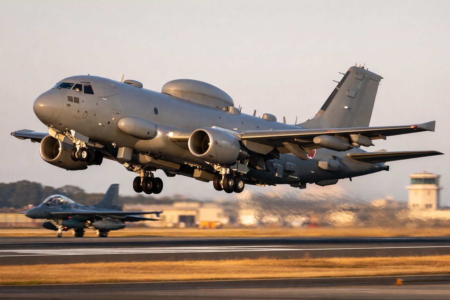 Avião militar cinzento a descolar de uma pista, com outro avião de caça estacionado ao fundo.