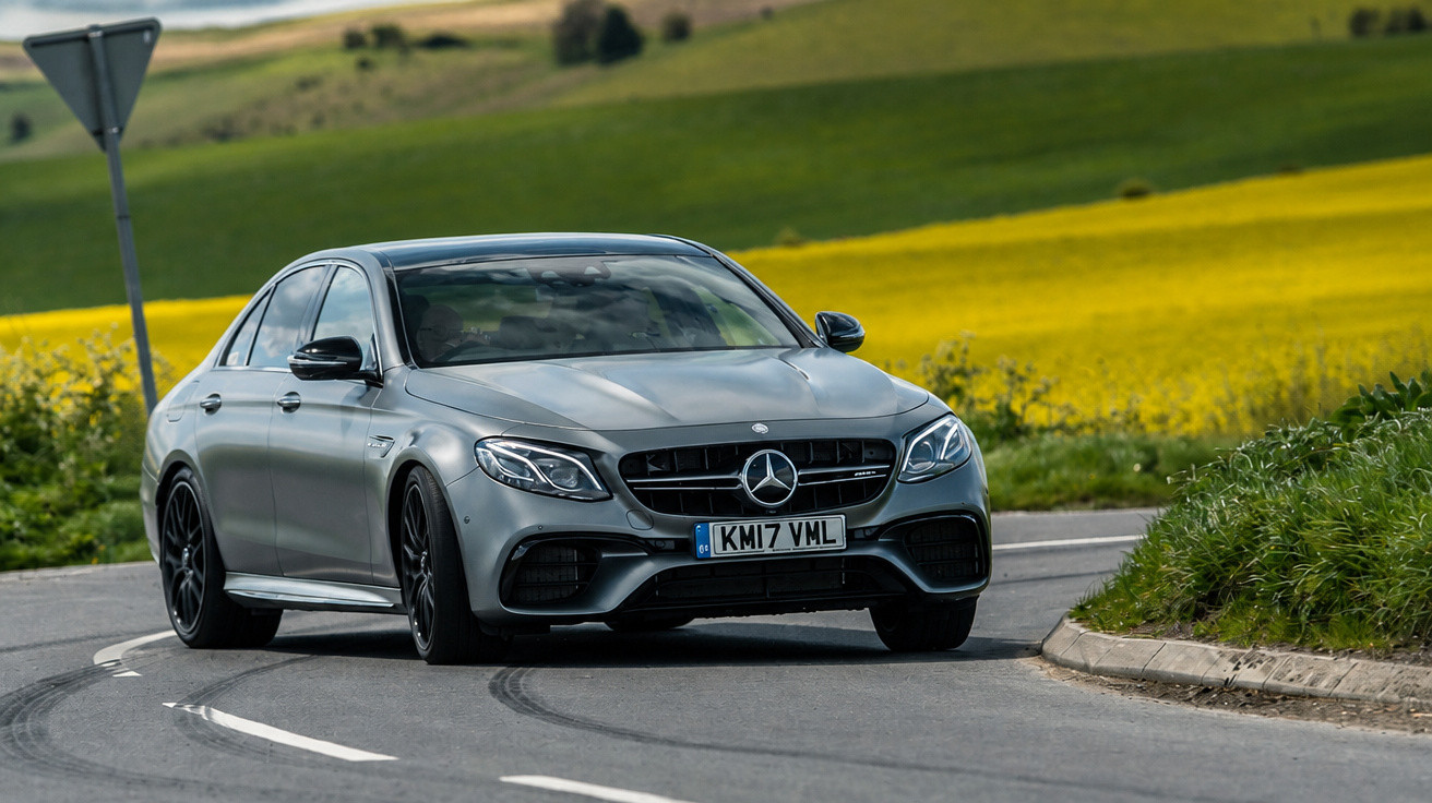 Carro Mercedes-Benz cinzento a contornar uma curva numa estrada rural com campos verdes e amarelos ao fundo.